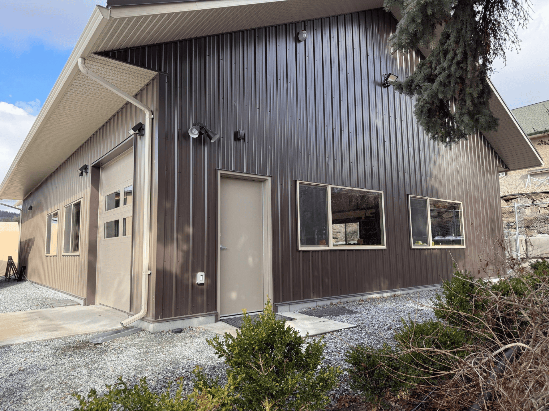 Exterior view of a custom woodworking shop with a modern metal facade, featuring large windows and a garage door, surrounded by gravel landscaping and greenery, representing Beaver Custom Builds' commitment to quality craftsmanship in woodworking services.